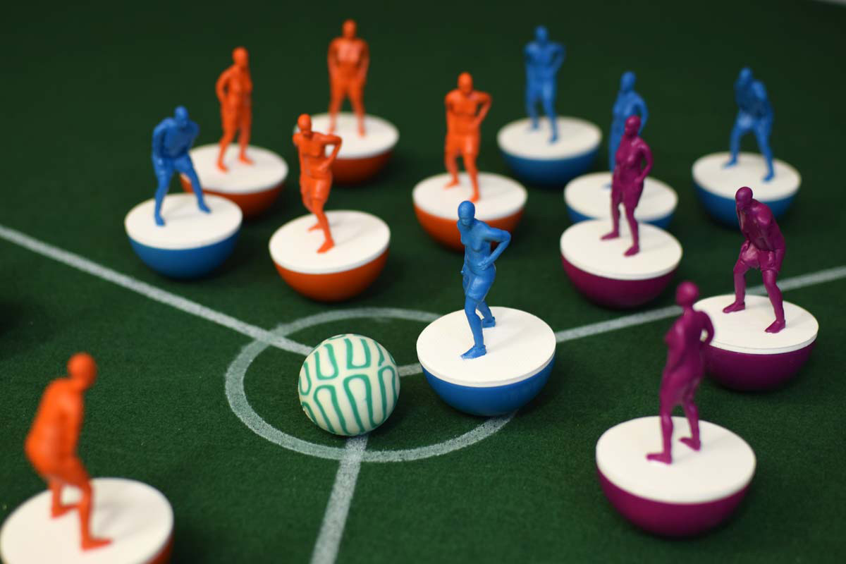 photo of subbuteo style women and non-binary 3d printed footballers, in red, turquoise and purple 5 of each colour on a green felt hexagon
