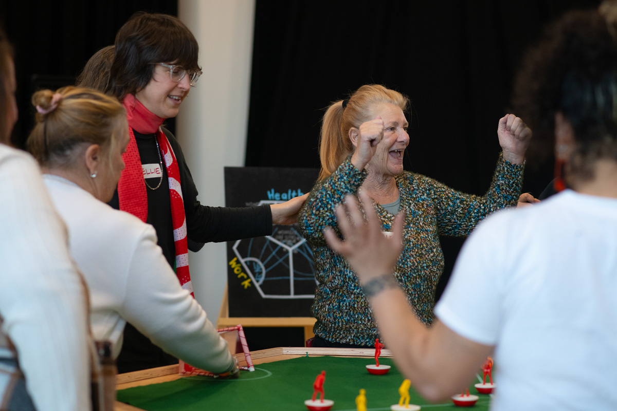 a group of older women playing feminist tabletop football with subbuteo-style women and non-binary 3d printed footballers on green felt